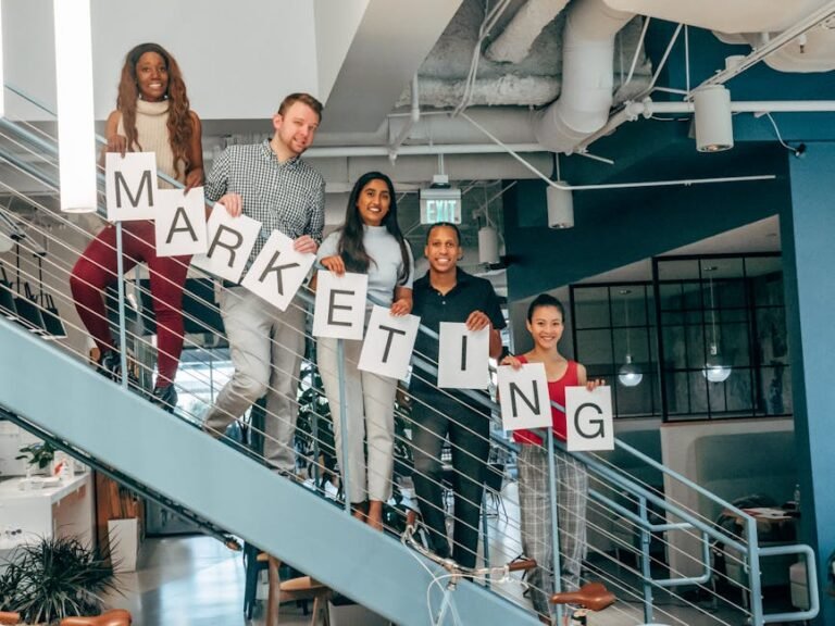 Five colleagues smiling and holding a marketing sign on an office staircase, showcasing teamwork.